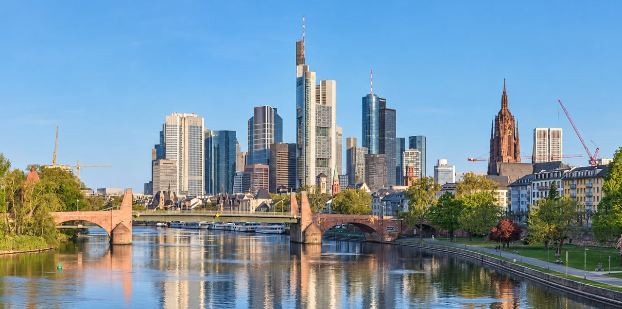 The River Main, Alte Bruecke and Frankfurt skyline beneath a clear blue sky, reflected in the water.